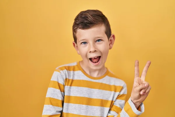 黄色の背景の上に立っている若い白人の子供は 勝利の兆候を行うカメラでの幸せな顔のウィンクで笑顔 — ストック写真