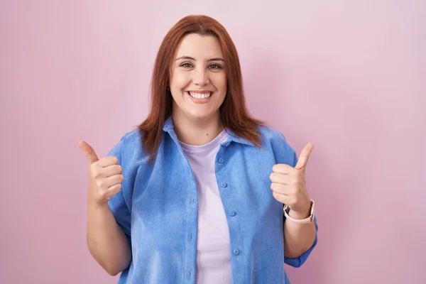 ピンクの背景の成功のサインの上に立っている赤い髪を持つ若いヒスパニック系の女性は 手で積極的なジェスチャーを行うと 親指を笑顔と幸せ 陽気な表情と勝者のジェスチャー — ストック写真