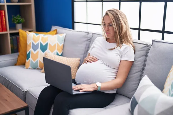 Young pregnant woman sitting on the sofa at home using laptop thinking attitude and sober expression looking self confident