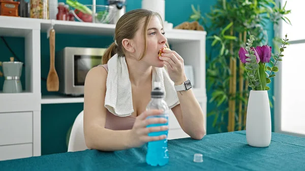 身穿运动服的金发年轻女子喝着精力充沛的饮料 在饭厅里吃苹果 — 图库照片