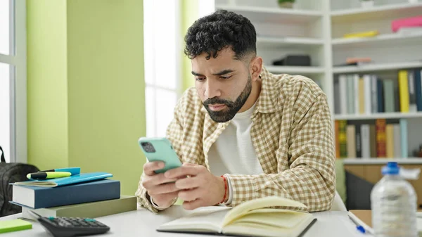 Young hispanic man student reading book using smartphone at library university