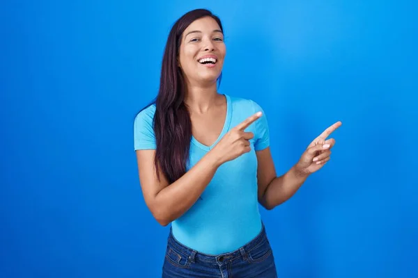 若いですヒスパニック系の女性が立っている青の背景笑顔とカメラを見て2本の手と指で側面を指して — ストック写真