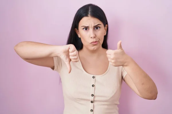 Young hispanic woman standing over pink background doing thumbs up and down, disagreement and agreement expression. crazy conflict