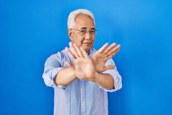 Hispanic senior man wearing glasses rejection expression crossing arms and palms doing negative sign, angry face