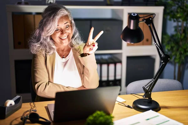 中年妇女白发苍苍 深夜用电脑笔记本电脑工作 脸上挂着喜色的笑容看着摄像机做胜利的标志 第二点 — 图库照片