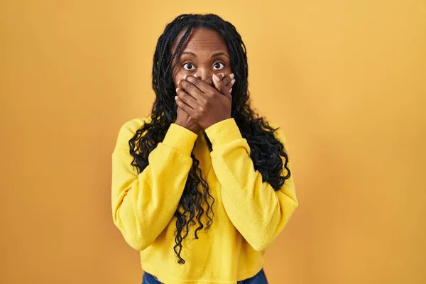 黄色の背景の上に立っているアフリカの女性は間違いのために手で口をカバーショックを受けた 秘密の概念 — ストック写真