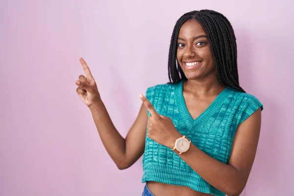 年轻的非洲裔美国人 头上戴着辫子 站在粉色的背景上笑着 用双手和手指看着摄像机的侧面 — 图库照片