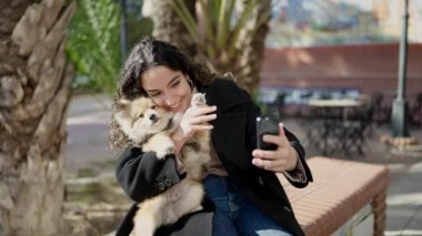 Köpeğiyle gülümseyen genç İspanyol kadın parkta akıllı telefondan selfie çekiyor.