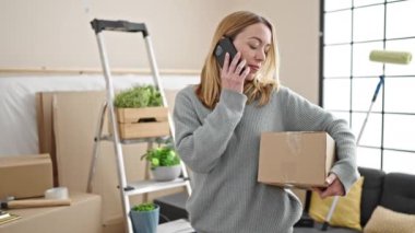 Genç sarışın kadın yeni evinde akıllı telefon paketiyle konuşuyor.