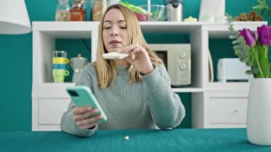 Genç sarışın kadın yemek odasında pirinç keki yerken akıllı telefon kullanıyor.