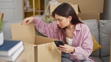 Young beautiful hispanic woman using smartphone unpacking cardboard box at new home