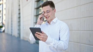 Young hispanic man talking on smartphone using touchpad at street
