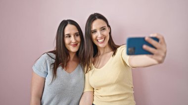 Kendinden emin gülümseyen iki kadın, izole pembe arka planda akıllı telefondan selfie çekiyor.