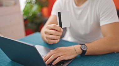 Laptop ve kredi kartıyla alışveriş yapan İspanyol genç adam yemek odasında masada oturuyor.