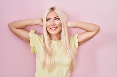 Beyaz kadın pembe arka planda duruyor rahatlıyor ve esniyor, kolları ve elleri başının ve boynunun arkasında mutlu bir şekilde gülümsüyor. 