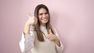 Genç, güzel, İspanyol bir kadın pembe arka planda baş parmağıyla gülümsüyor.
