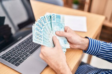 Young man business worker using laptop holding brazilian real banknotes at office