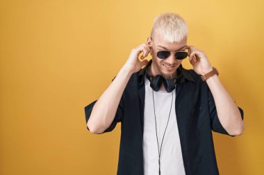 Sarı arka planda duran güneş gözlüğü takan genç beyaz adam yüksek sesli müzik sesi için kulaklarını parmaklarıyla kapatıyor. sağır kavramı. 