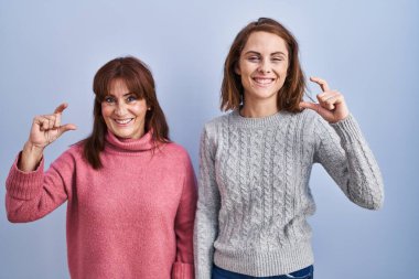 Anne ve kızı mavi arka planda durup gülümsüyor ve el hareketleriyle el hareketleriyle küçük boy işareti yapıyor ve kameraya bakıyor. ölçüm kavramı. 