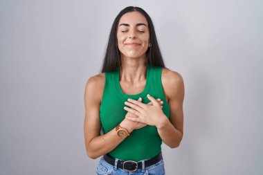 Soyutlanmış arka planda duran genç bir kadın elleri göğsünde, gözleri kapalı ve yüzünde minnet dolu bir jest ile gülümsüyor. sağlık kavramı. 