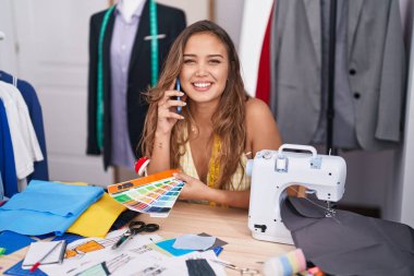 Young beautiful hispanic woman tailor talking on smartphone looking color test at tailor shop