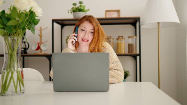 Genç kızıl saçlı kadın dizüstü bilgisayar kullanıyor ve evdeki akıllı telefondan konuşuyor.