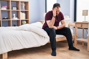 Young hispanic man sitting on bed with sad expression at bedroom