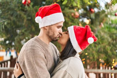 Erkek ve kadın çift Noel şapkası takıyor. Birbirlerine sarılıyorlar ve parkta öpüşüyorlar.