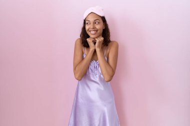 Genç İspanyol kadın uyku maskesi takıyor ve geceliği var. Gergin ve heyecanlı bir şekilde gülüyor, elleri çenesinde yan tarafa bakıyor. 