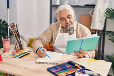 Sanat stüdyosunda kitap okuyan orta yaşlı, kır saçlı bir sanatçı.