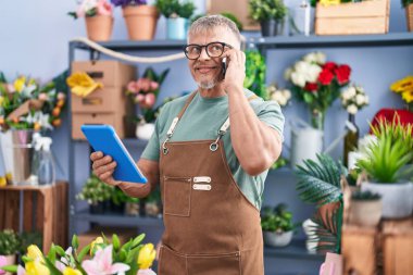 Orta yaşlı, kır saçlı çiçekçi. Çiçekçide akıllı telefondan konuşan dokunmatik ped kullanıyor.