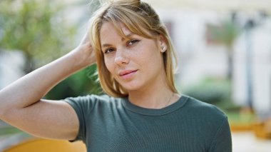 Genç sarışın kadın ciddi bir ifadeyle parkta saçını tarıyor.