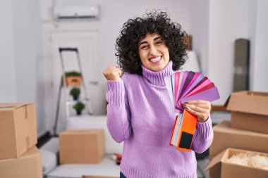 Yeni ev duvarının rengini seçen kıvırcık saçlı genç esmer kadın gururla bağırıyor, zaferi kutluyor ve kollarını kaldırarak başarıyı kutluyor. 