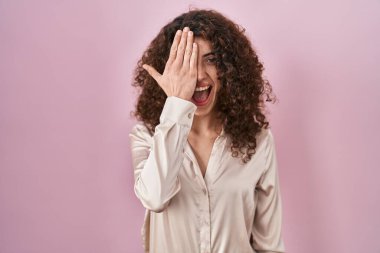 Hispanic woman with curly hair standing over pink background covering one eye with hand, confident smile on face and surprise emotion. 