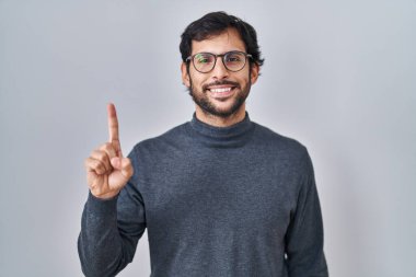 Yakışıklı Latin adam izole edilmiş arka planda duruyor. Bir numarayı gösterip işaret ederken kendine güvenen ve mutlu bir şekilde gülümsüyor.. 