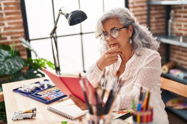 Sanat stüdyosunda kitap okuyan orta yaşlı bir kadın sanatçı.