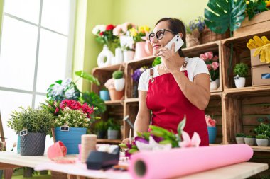 Young beautiful hispanic woman florist talking on smartphone writing on notebook at flower shop