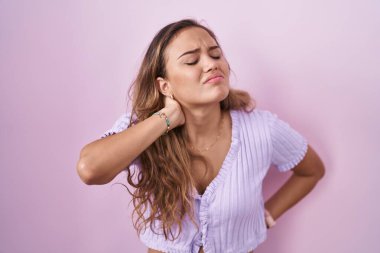 Young hispanic woman standing over pink background suffering of neck ache injury, touching neck with hand, muscular pain 