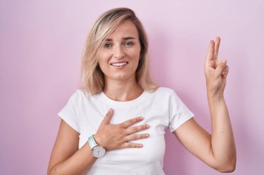 Young blonde woman standing over pink background smiling swearing with hand on chest and fingers up, making a loyalty promise oath 