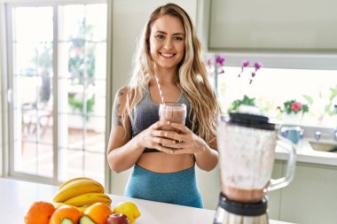 Mutfakta elinde bir bardak meyve püresi tutarak gülümseyen genç bir kadın.
