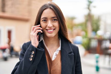 Genç, güzel, İspanyol bir kadın gülümsüyor ve caddede akıllı telefondan konuşuyor.