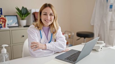 Genç ve güzel İspanyol doktor dizüstü bilgisayar kullanıyor. Masada oturmuş klinikte kollarını kavuşturup el kol hareketi yapıyor.