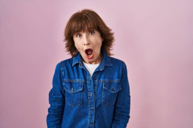 Middle age woman standing over pink background afraid and shocked with surprise and amazed expression, fear and excited face. 