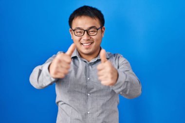 Mavi arka planda duran genç Çinli adam elleriyle olumlu jest yapmayı onaylıyor, gülümsüyor ve başarı için mutlu. kazanan jest. 