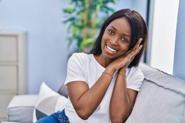 Genç Afrikalı Amerikalı kadın gülümsüyor. Kendine güvenen, evinde elleriyle uyuyan jest yapan.