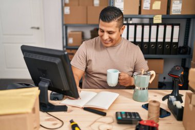 Young latin man ecommerce business worker drinking coffee writing on notebook at office
