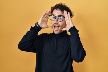 Hispanic man standing over yellow background smiling cheerful playing peek a boo with hands showing face. surprised and exited 