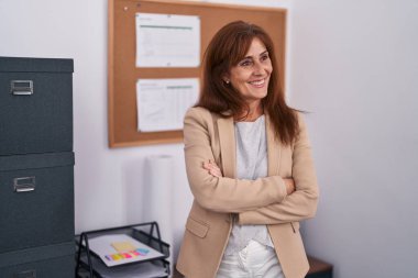 Orta yaşlı iş kadını güler yüzlü, kendine güler yüzlü, kollarını kavuşturup ofiste el kol kola duran biri.