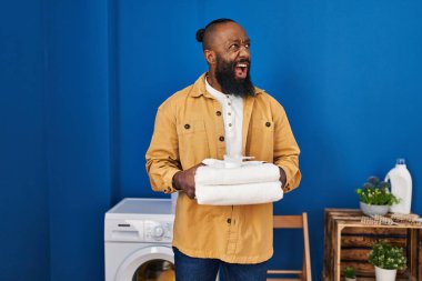 Afrika kökenli Amerikalı adam çamaşır odasında temiz havlu tutuyor kızgın ve öfkeli çığlıklar atıyor öfkeyle bağırıyor. öfke ve agresif konsept. 