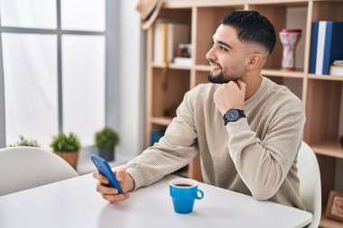 Young arab man using smartphone and drinking coffee sitting on table at home